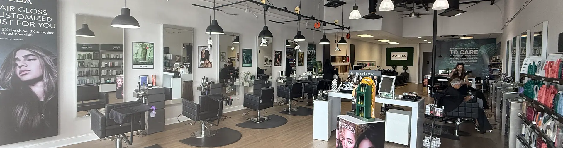 Modern hair salon interior with empty styling chairs, mirrors, product displays, and a few people seated; bright lighting and minimalist decor.