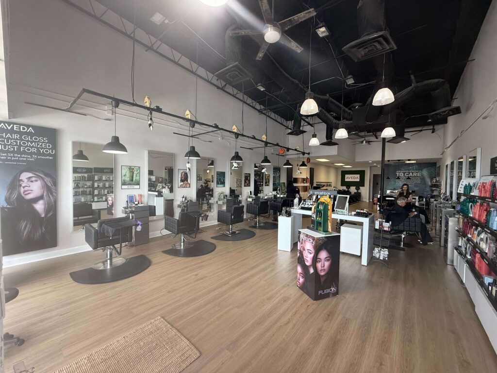 Modern hair salon interior with styling chairs, mirrors, hair products on shelves, and two people seated near the back reception area. The space has high ceilings and wood flooring.