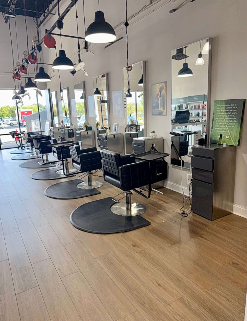 A modern hair salon with black styling chairs, large mirrors, and wooden flooring, with natural light coming in through large windows.