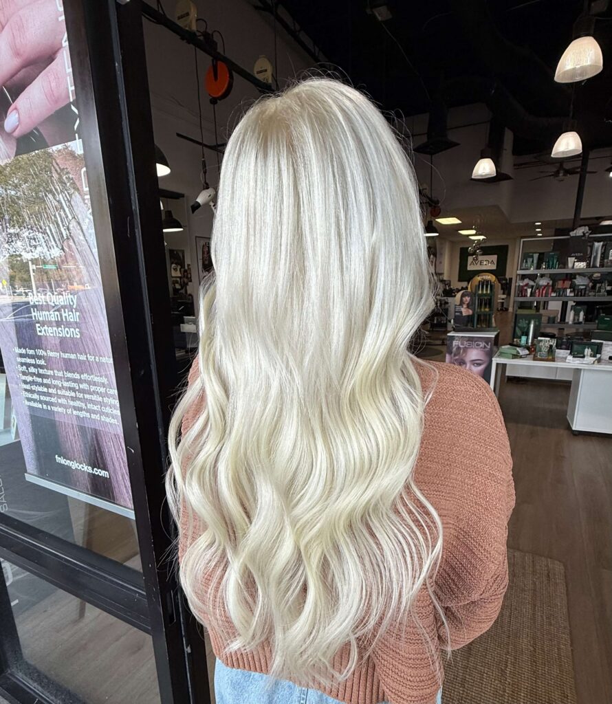 A person with long, wavy, platinum blonde hair is standing indoors, facing away from the camera. They are wearing a brown sweater and light blue jeans.