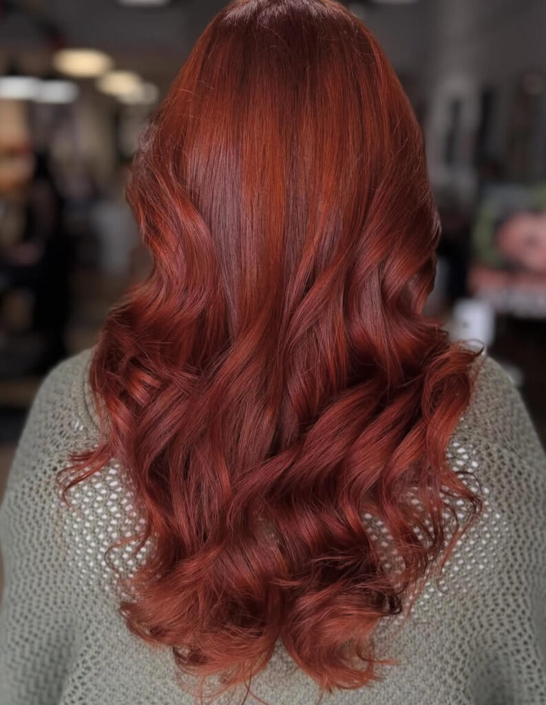 Person with long, wavy, reddish-brown hair seen from behind, wearing a light knit sweater in an indoor setting.
