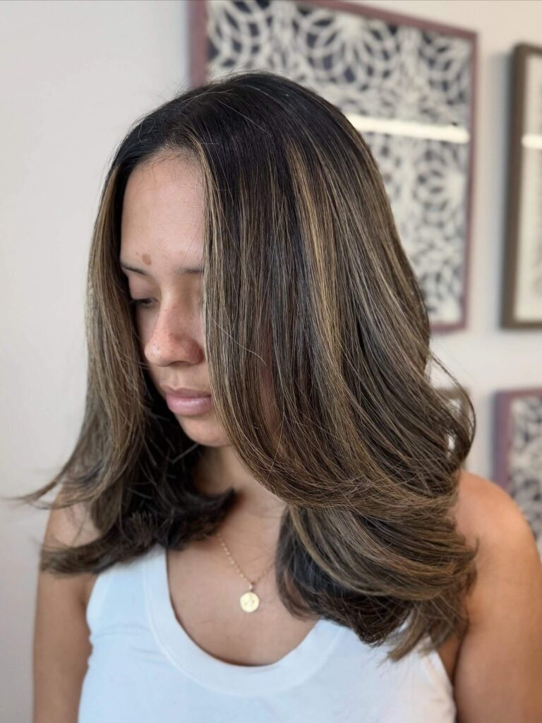 Woman with shoulder-length, layered brown hair with blonde highlights, wearing a white tank top and a gold necklace, looking down in front of a patterned background.