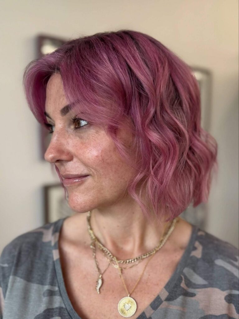 A woman with wavy, chin-length pink hair looks to the side, wearing a gray camouflage top and layered gold necklaces.
