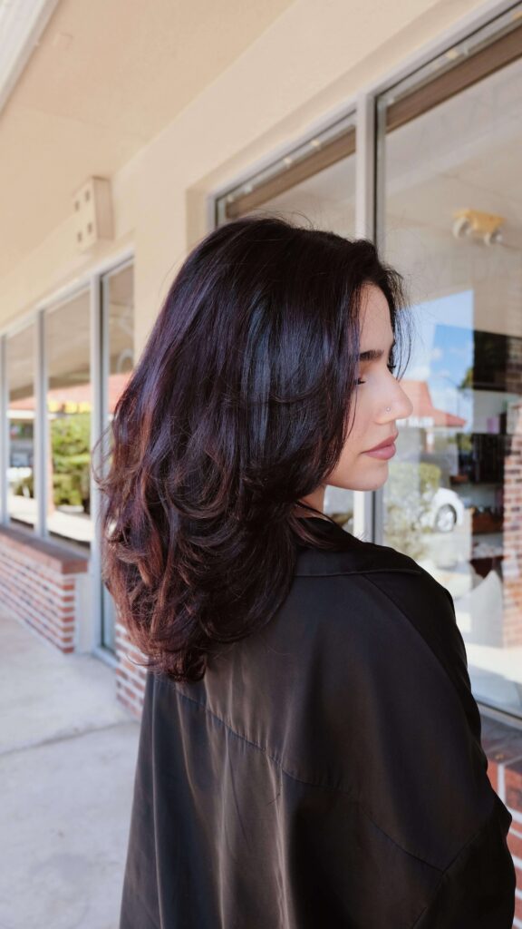 Woman with shoulder-length dark layered hair stands outside a building with large windows, facing slightly away from the camera.
