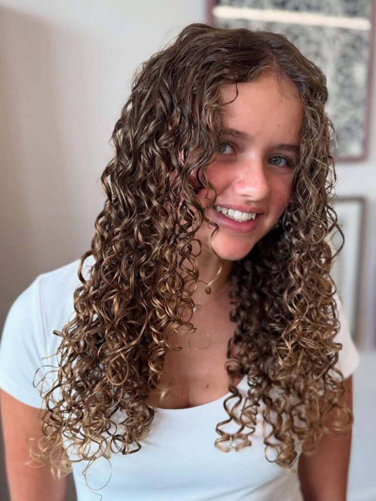 A young woman with long, defined brown curls smiles at the camera, wearing a white top and standing indoors against a blurred background.