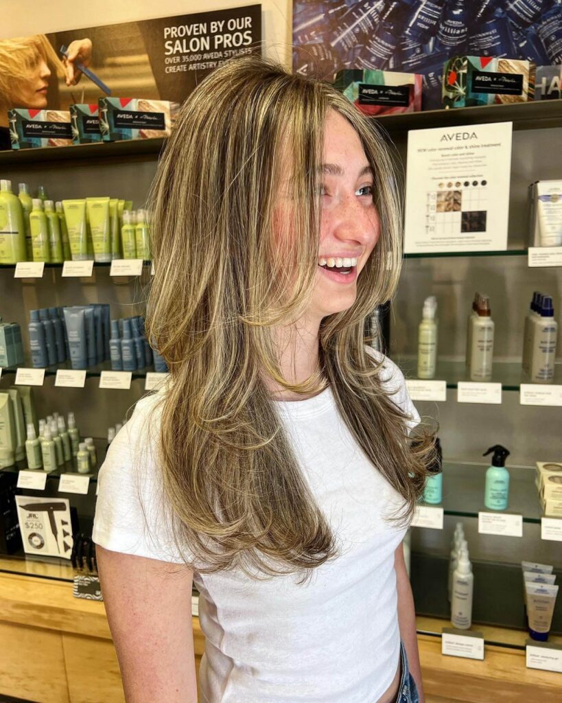 Woman with long, layered, blonde-highlighted hair wearing a white t-shirt, standing in a salon with hair products displayed on shelves behind her.