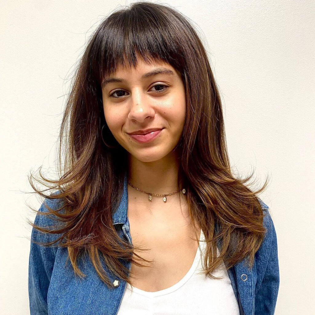 A young woman with layered brown hair and short bangs wears a denim jacket over a white top, standing against a plain white background.
