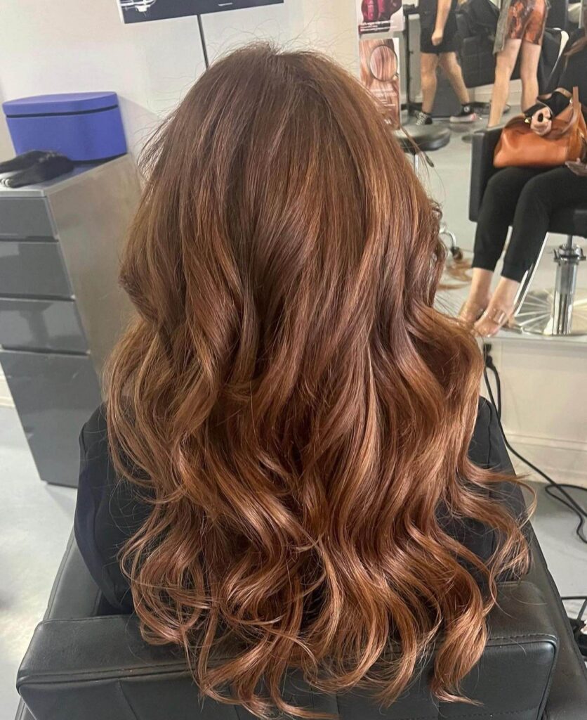 Person with long, wavy, light brown hair sits in a salon chair facing away from the camera; salon equipment and another seated person are visible in the background.