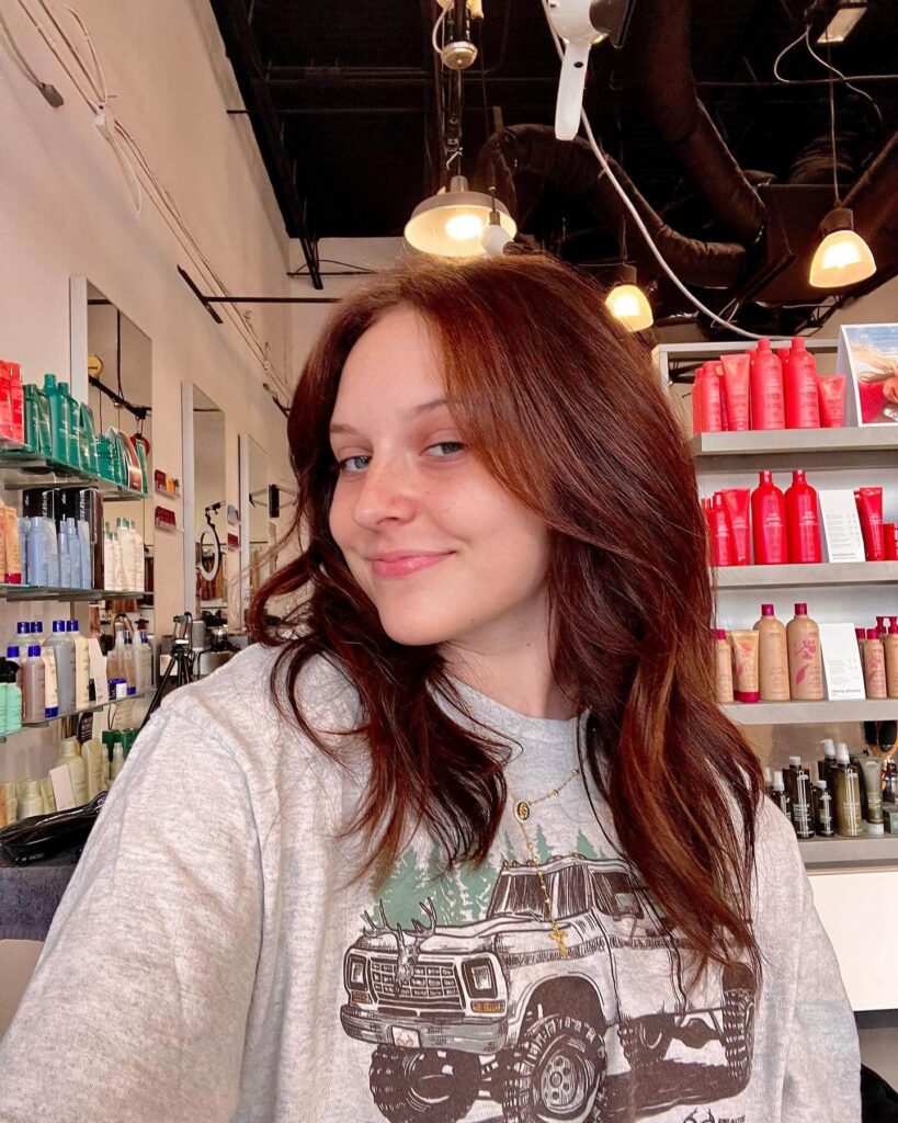 A woman with medium-length brown hair smiles slightly, wearing a grey shirt with a truck graphic, inside a salon with shelves of hair products behind her.