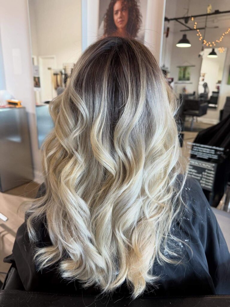 A person with long, wavy, blonde balayage hair sits in a salon chair, facing away from the camera. Salon mirrors, lights, and posters are visible in the background.