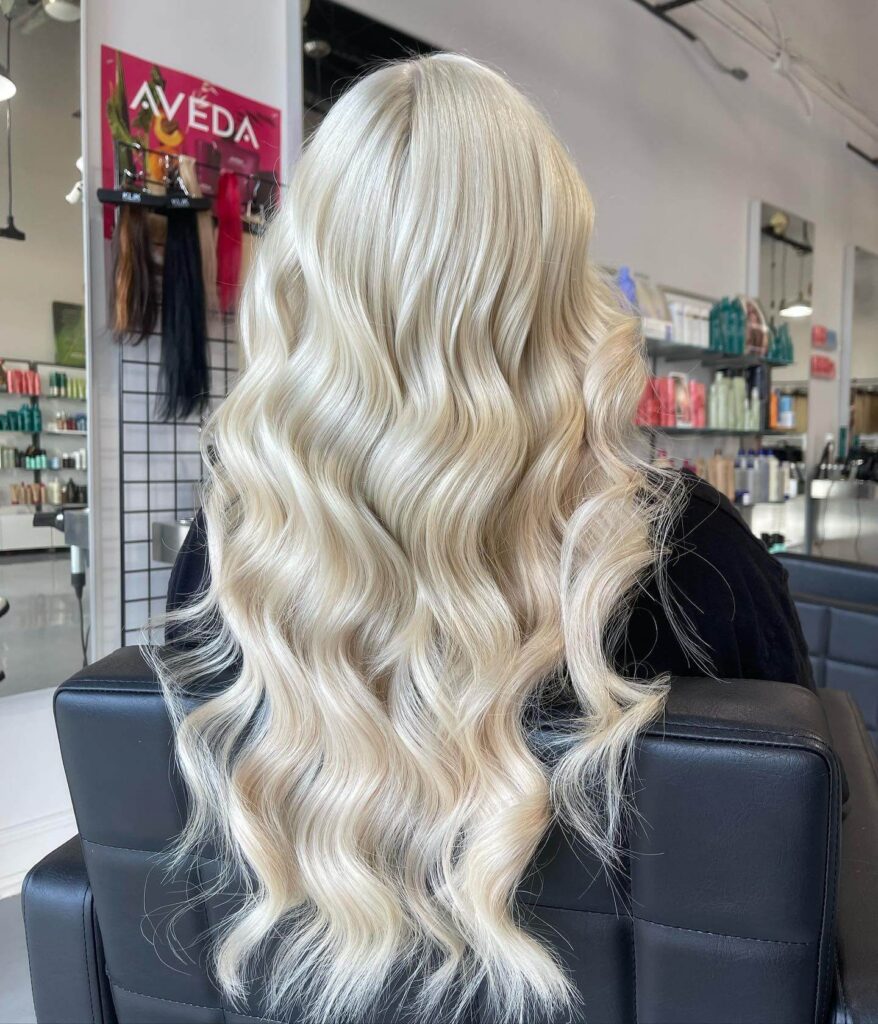 A person with long, platinum blonde, wavy hair sits in a salon chair facing away, with hair products and salon tools visible in the background.