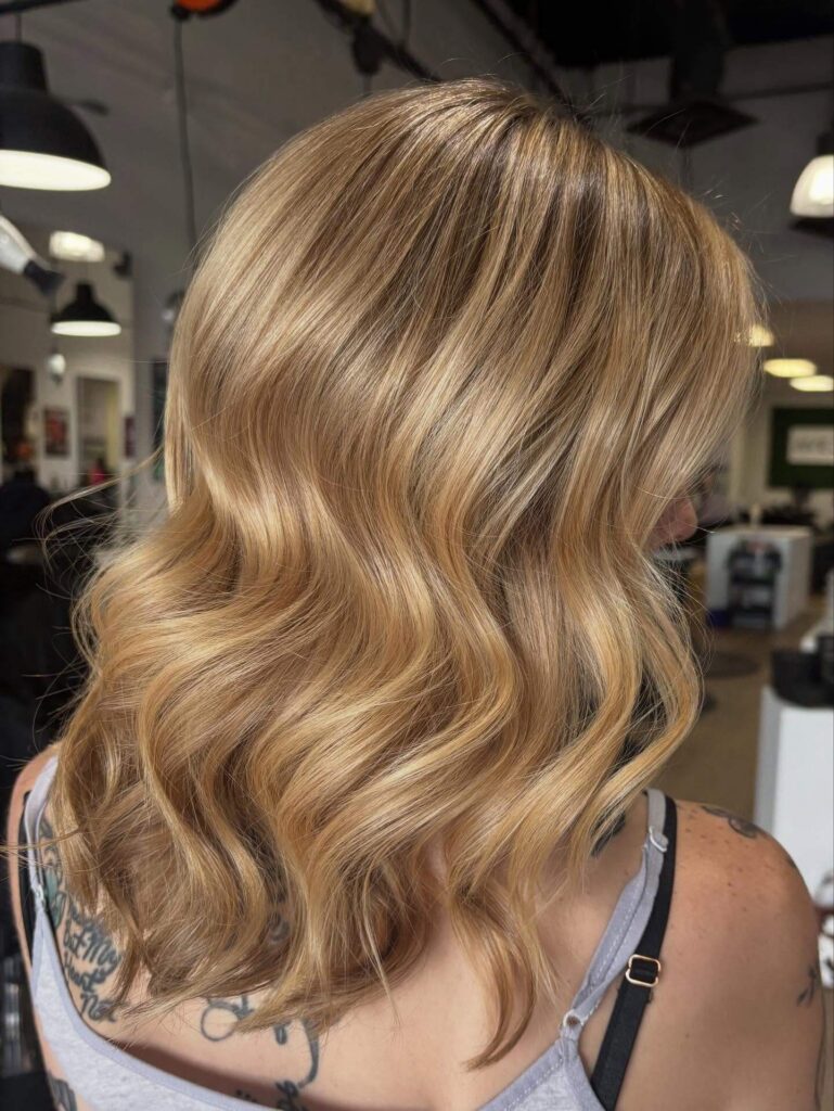 A person with wavy, shoulder-length blonde hair is pictured from behind in a well-lit salon. Tattoos are visible on their shoulders.