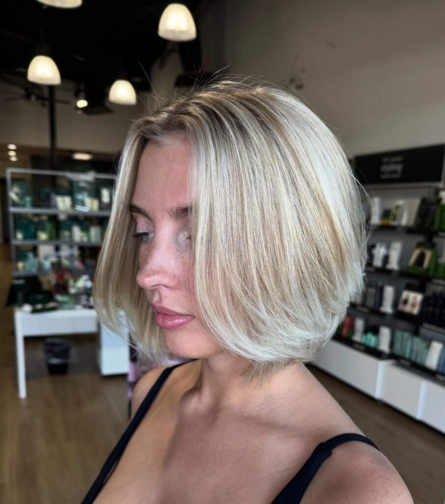 A woman with a blonde bob haircut and black sleeveless top stands in a modern hair salon with shelves of hair products in the background.